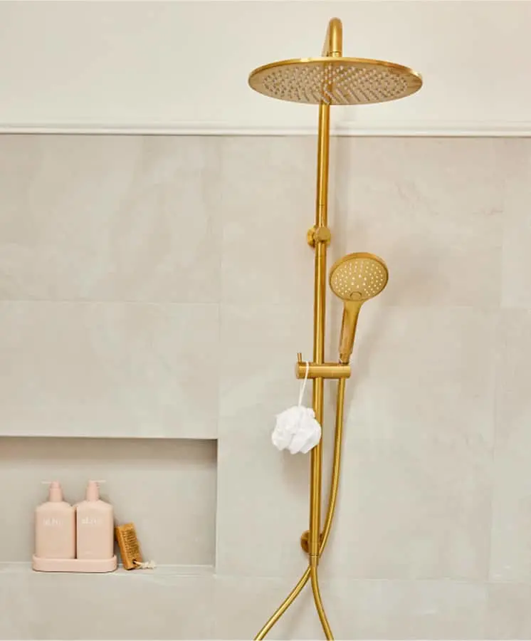 Gold rainfall shower system with handheld attachment mounted on light beige tiled wall with built-in shelf holding bath products.