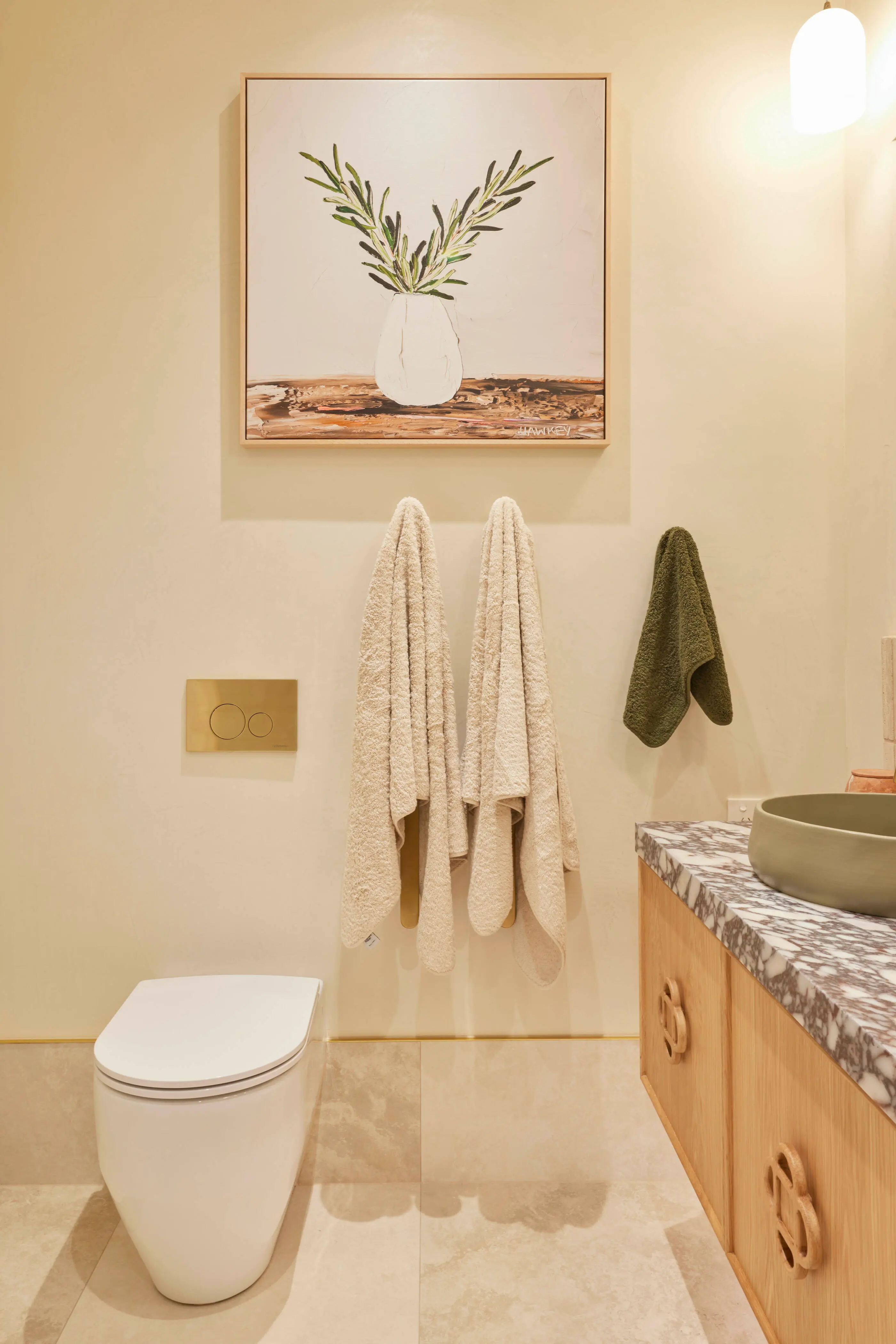 Modern bathroom with white toilet, wooden vanity, cream and green towels, and olive branch painting on beige wall.