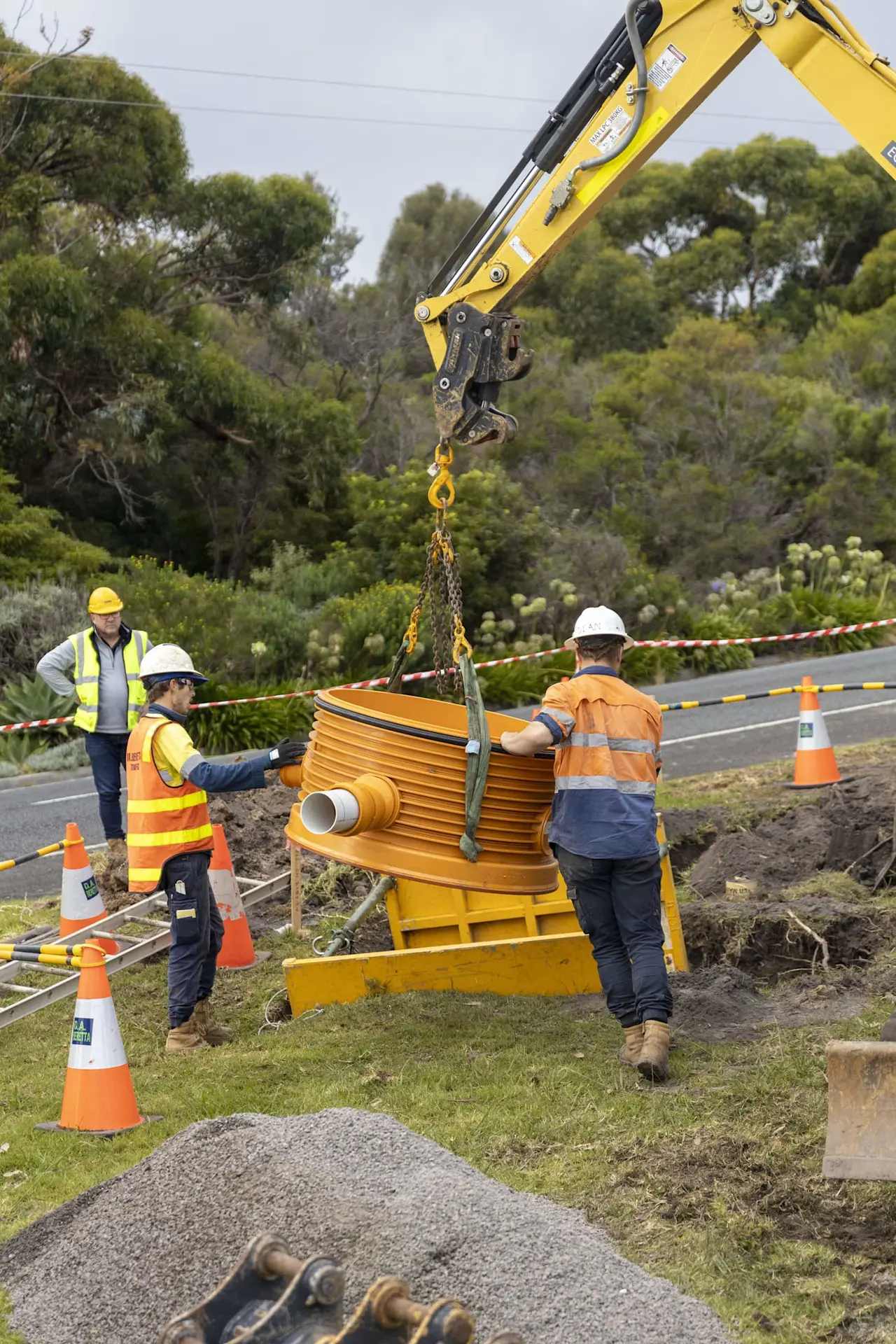 Construction workers installing yellow drainage equipment with a crane at roadside work site surrounded by safety cones.