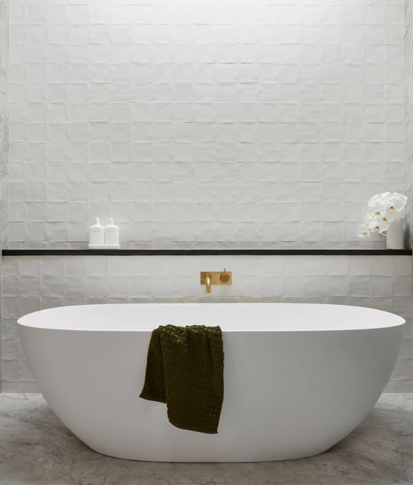 Modern white freestanding bathtub with dark green towel against textured white tile wall with gold fixtures and shelf.