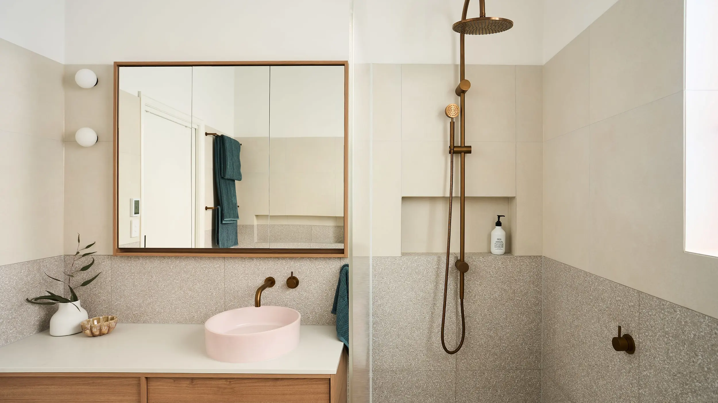 Modern bathroom with pink basin, wooden-framed mirror, brass fixtures, and walk-in shower with terrazzo-style tiling.