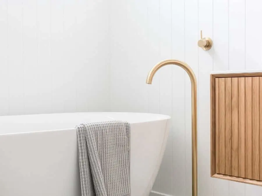 Modern white bathtub with gray towel, brass faucet, and wooden cabinet in a minimalist bathroom with white walls.