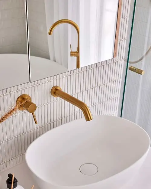 Modern bathroom with white oval sink, brass wall-mounted faucet, and vertical tile backsplash reflected in mirror.