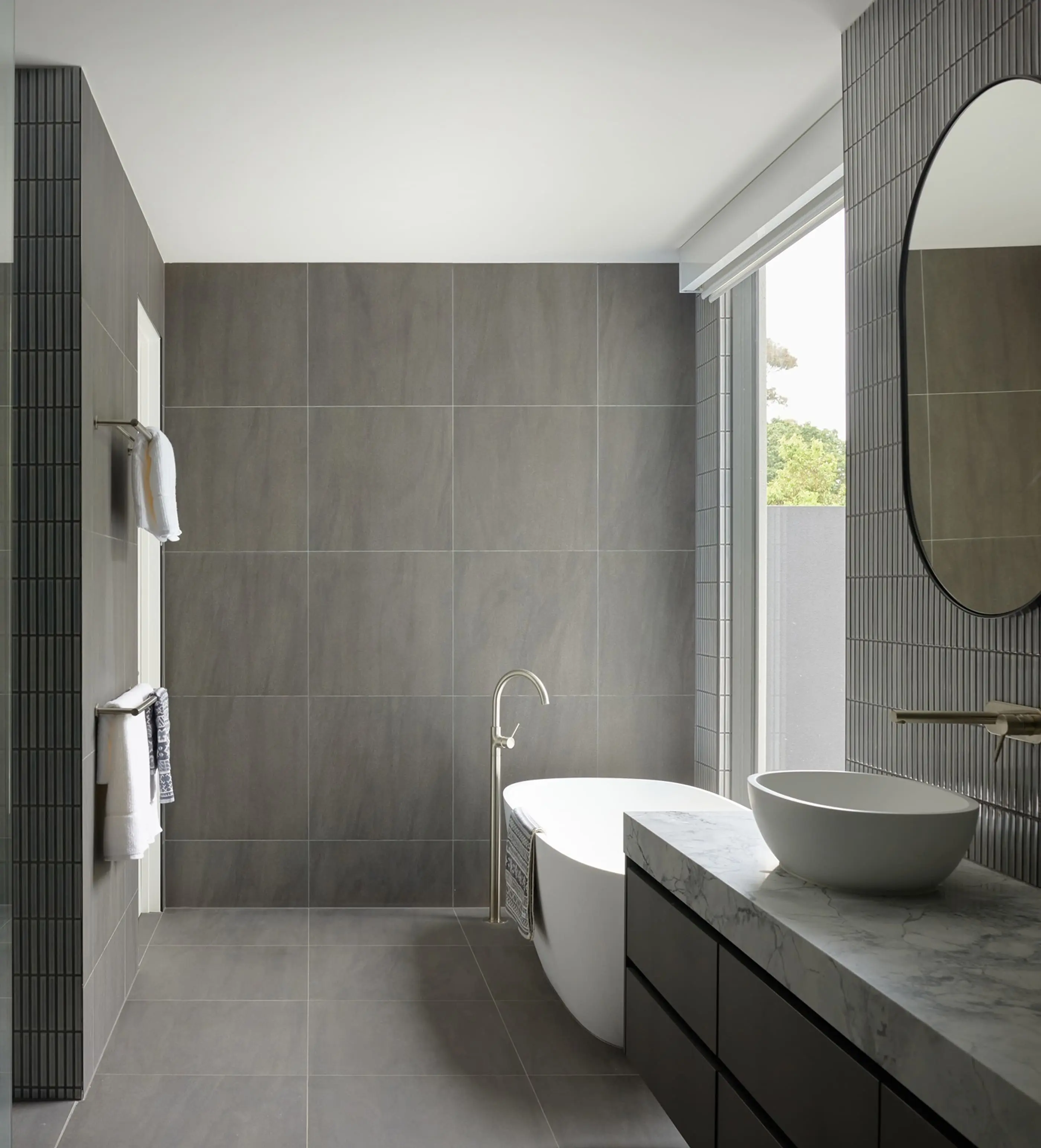 Modern bathroom with gray tile walls, white freestanding tub, vessel sink on marble countertop, and round mirror.