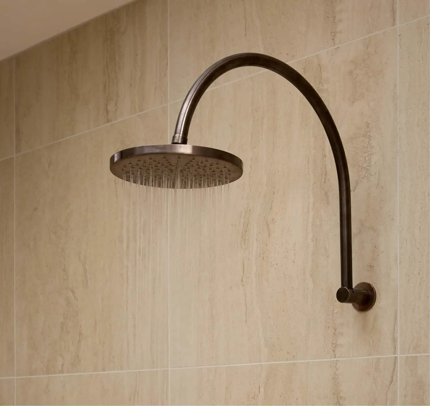 Bronze rainfall showerhead with curved arm mounted on beige tiled wall with water flowing.
