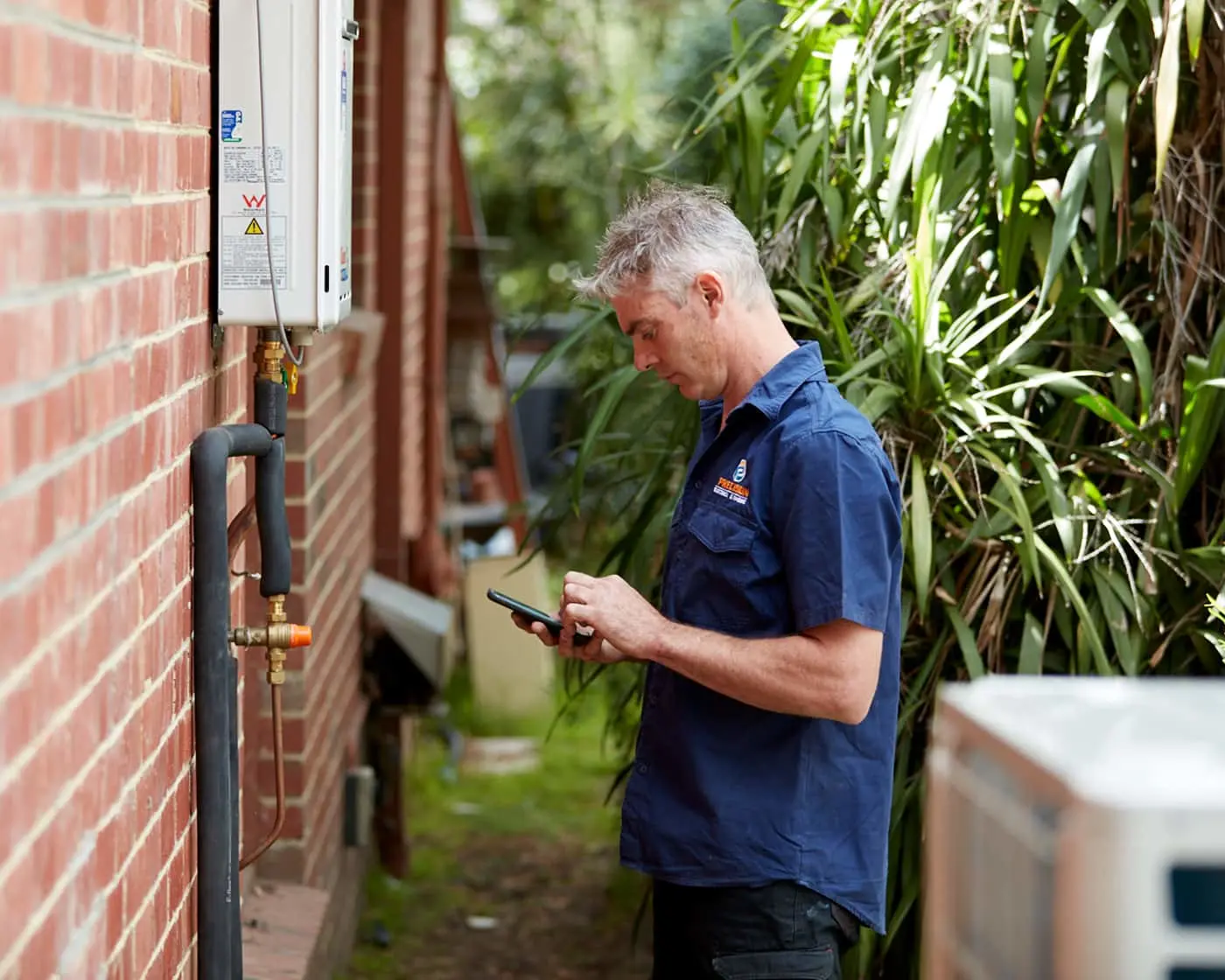 Technician in blue uniform checking phone while inspecting water heater mounted on brick wall exterior.