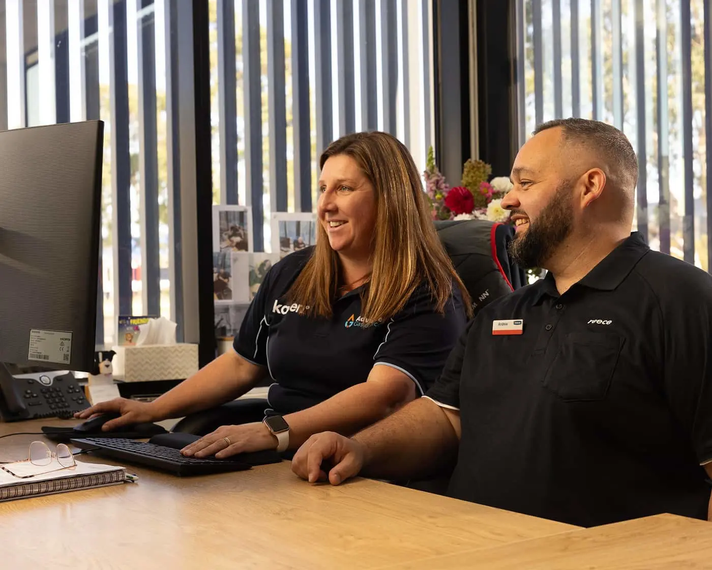 Two colleagues in black polo shirts smiling while working at a computer in an office with vertical blinds.