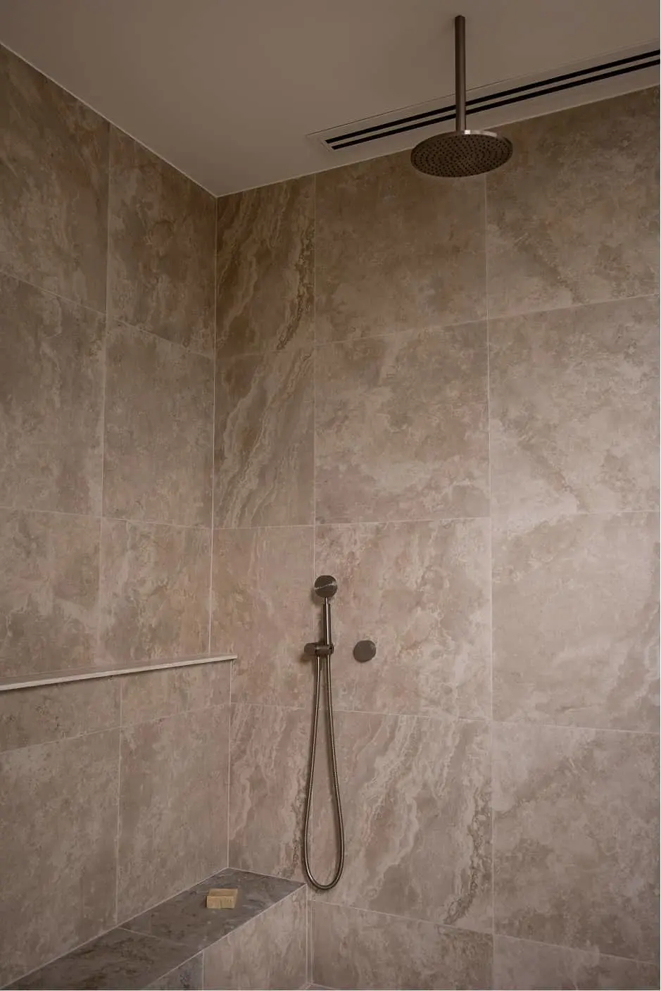 Modern shower with beige stone tiles, bronze rainfall showerhead, handheld sprayer, and built-in corner shelf.