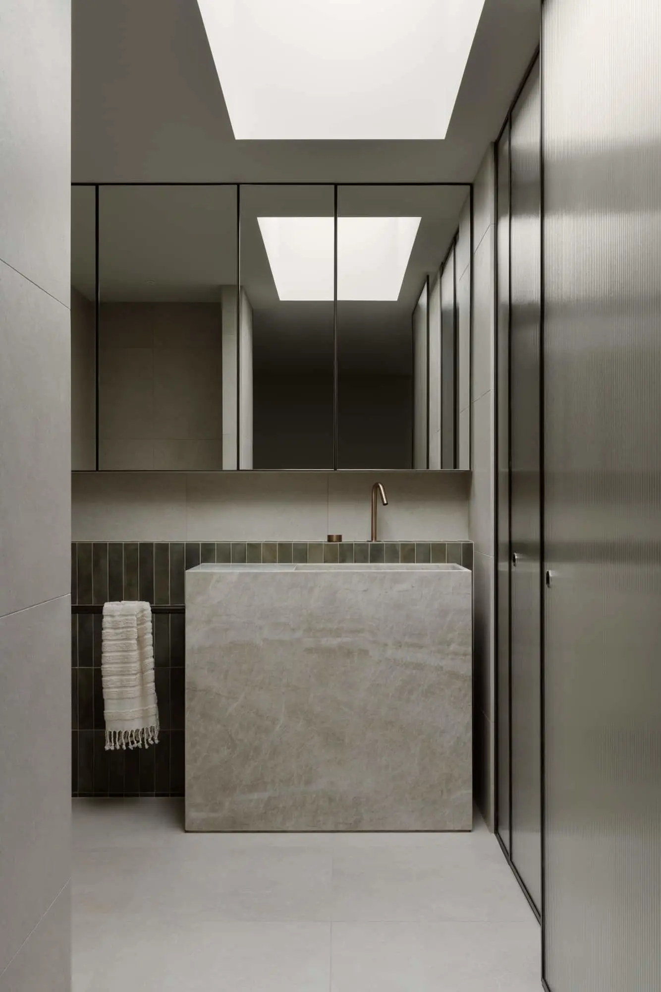 Modern minimalist bathroom with stone sink, mirrored cabinets, skylights, and textured glass partition.