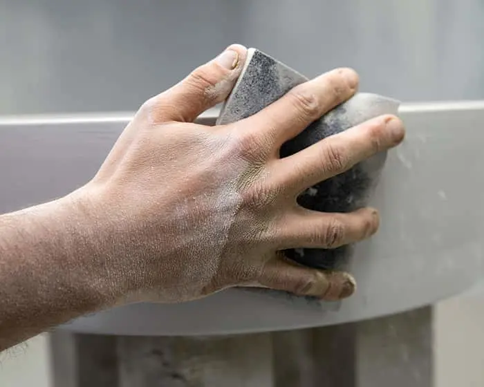Hand using a sanding block on a light-colored wall, with dust visible on the skin and surface.