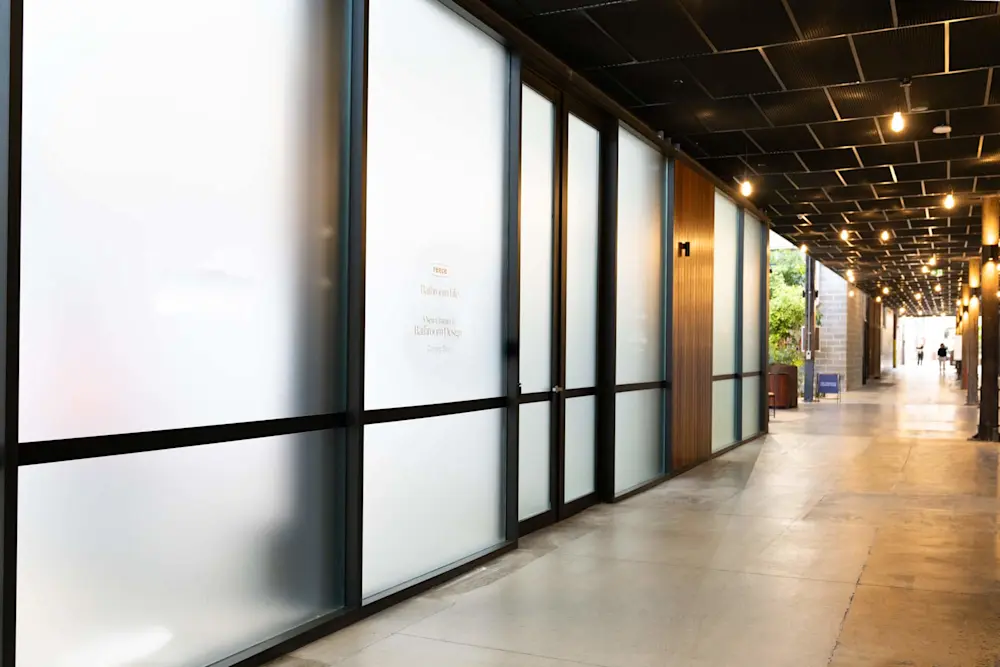 Modern corridor with frosted glass walls, pendant lighting, and wood accents leading to an outdoor area.