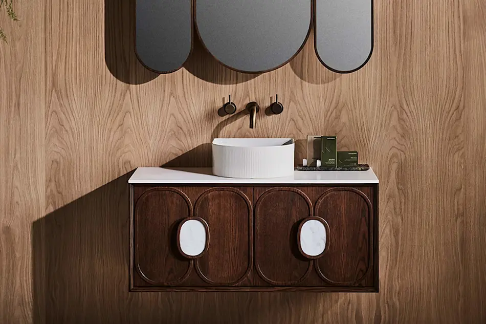 Modern bathroom vanity with wooden cabinet, white vessel sink, brass wall-mounted faucet, and oval mirrors on wood-paneled wall.