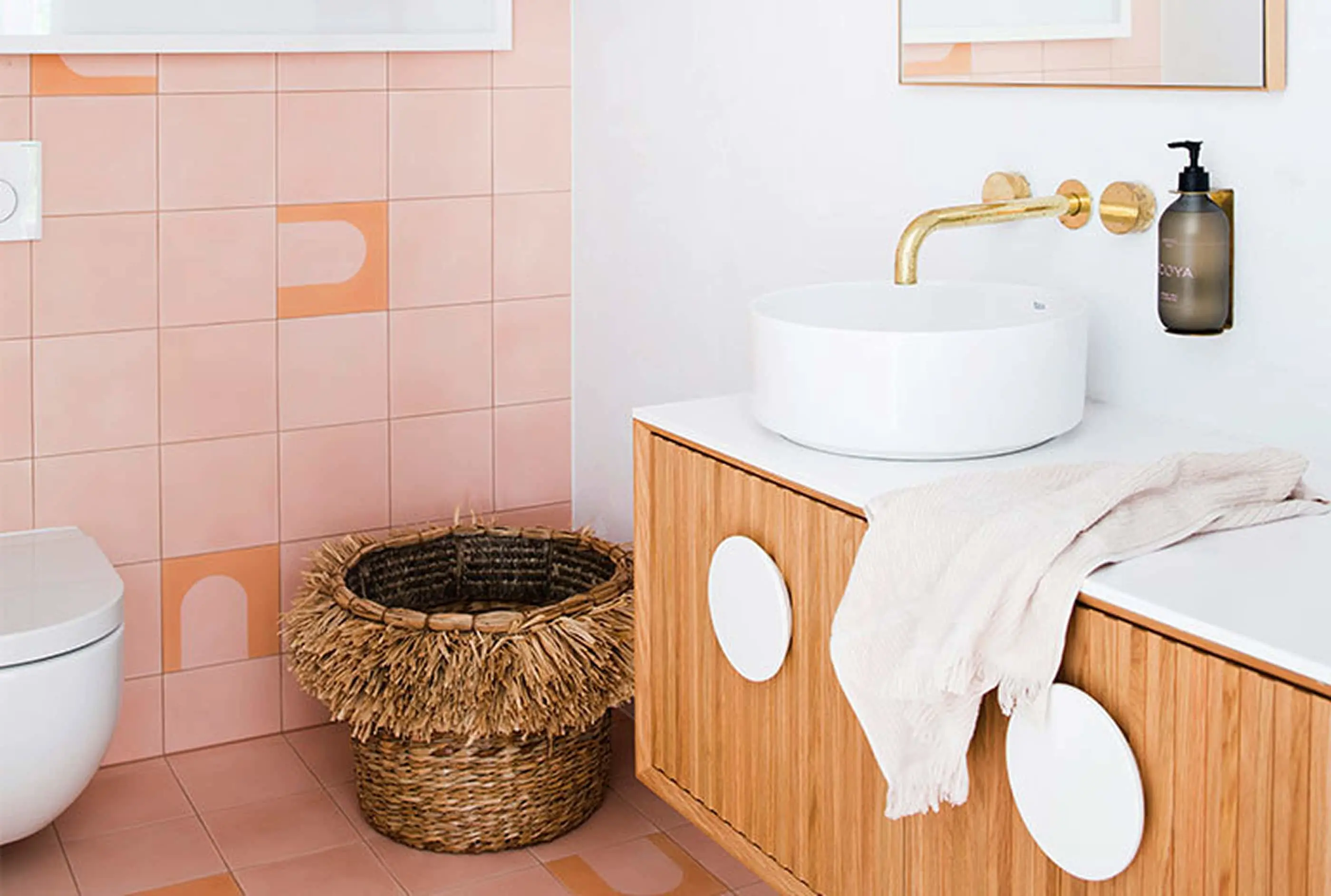 Modern bathroom with pink tiles, wooden vanity, white vessel sink, brass faucet, woven basket, and white towel.