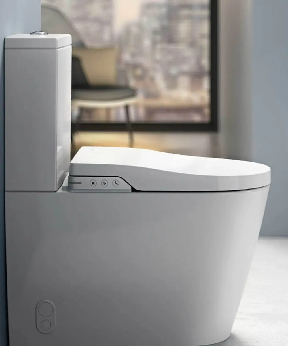 Modern white toilet with electronic bidet seat featuring control buttons, in a minimalist bathroom with blurred window view.