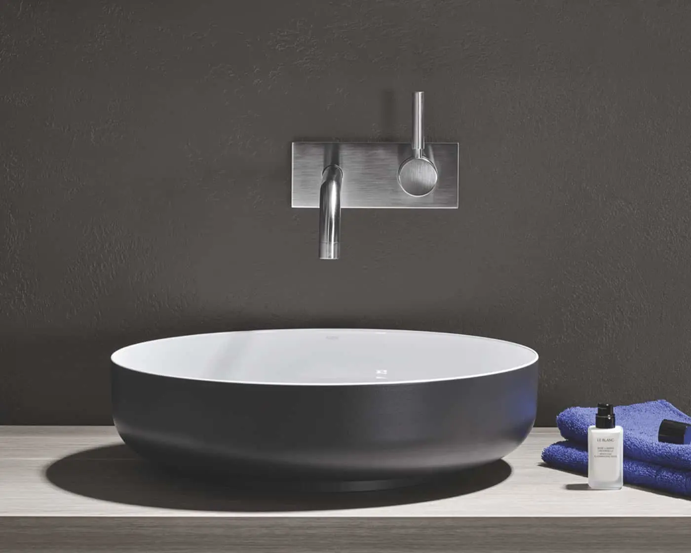 Modern bathroom sink with black exterior, white interior, wall-mounted steel faucet, blue towels and toiletry bottle.