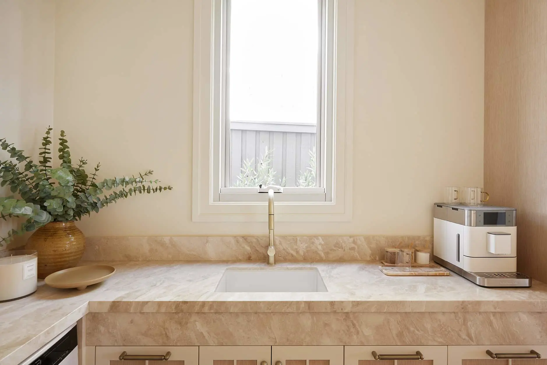 Modern kitchen with marble countertop, gold faucet, coffee machine, and eucalyptus plant in ceramic pot beneath window.