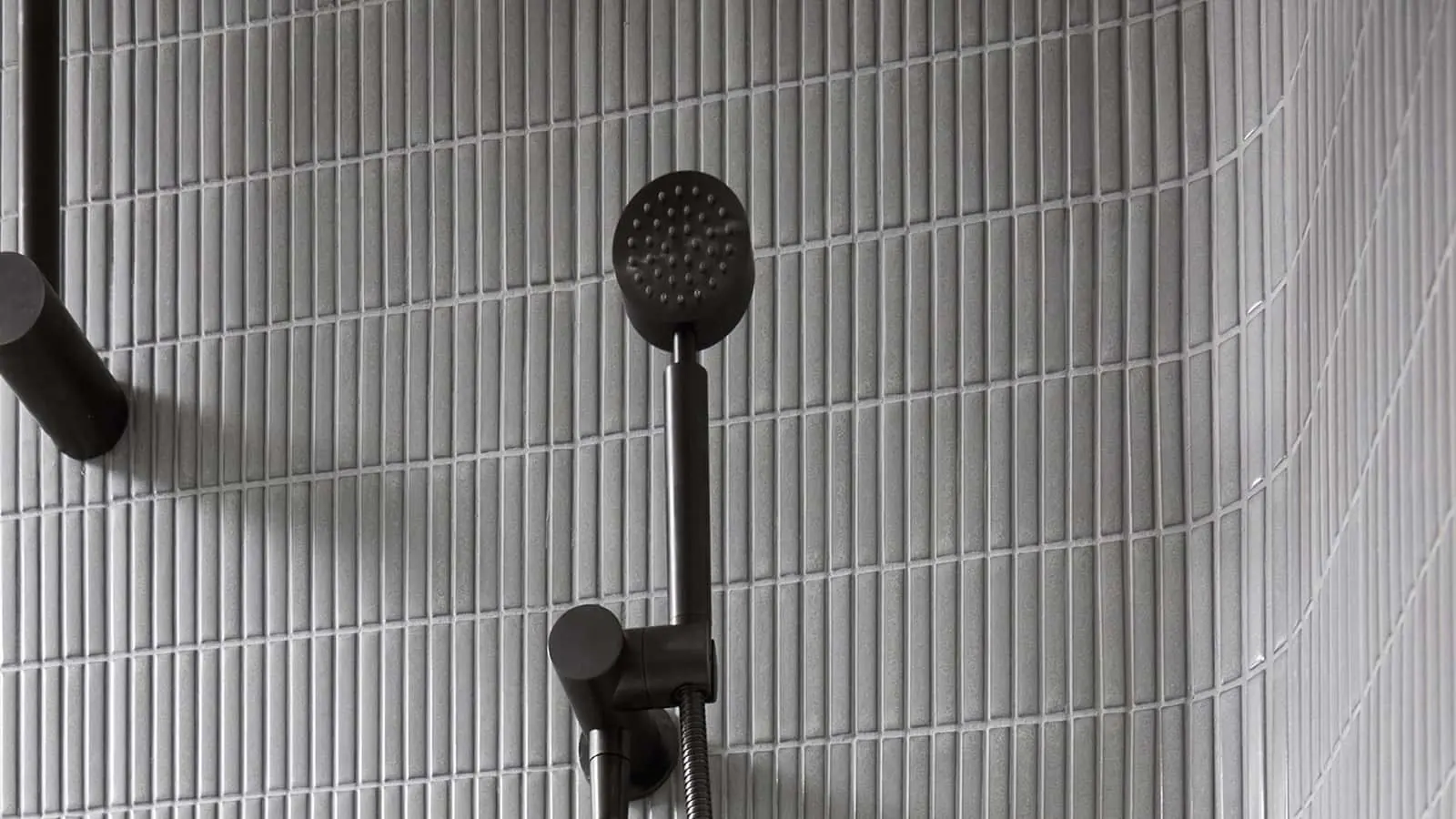 Black handheld shower head mounted on a curved wall with light gray vertical tile in a modern bathroom.