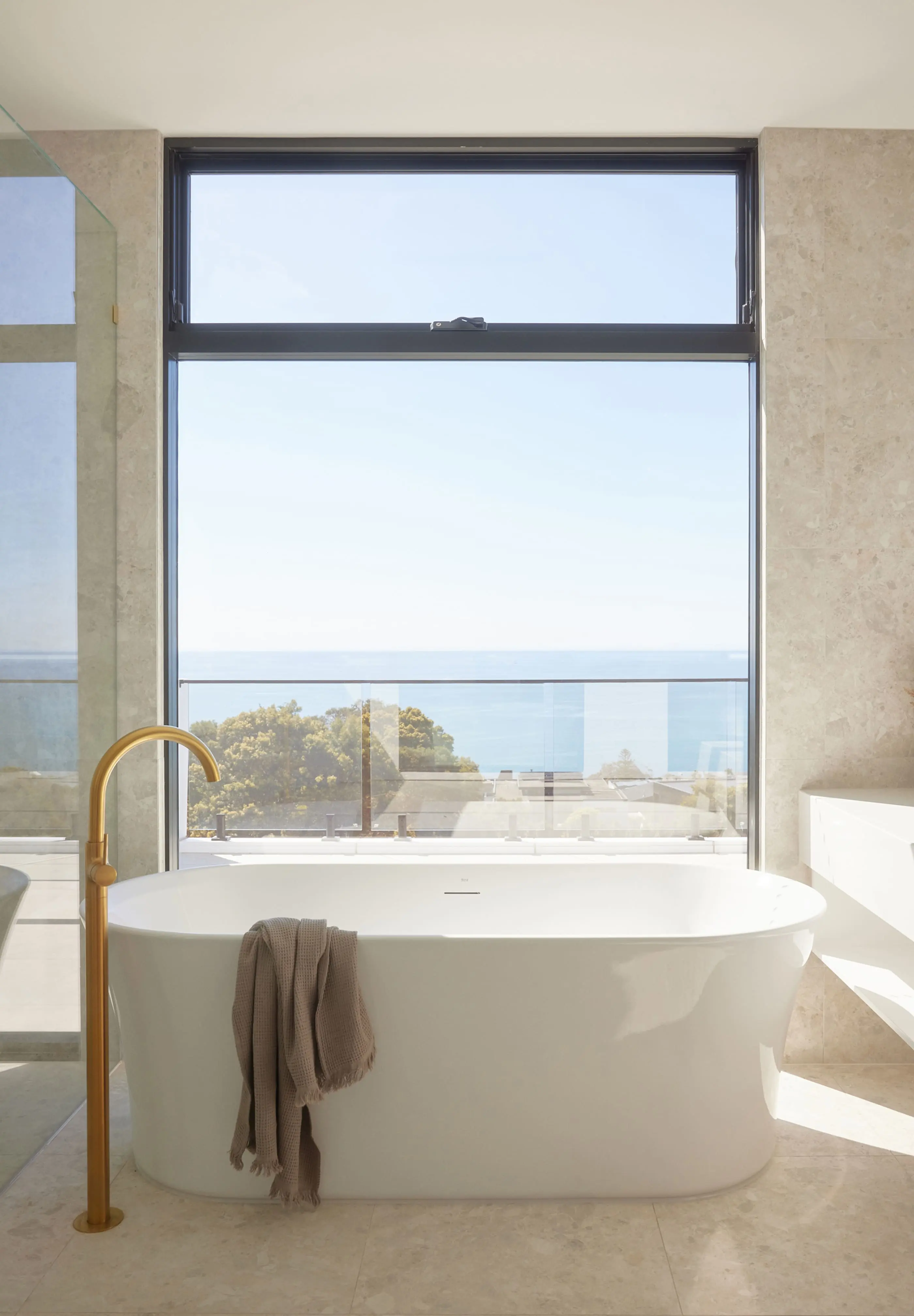 Modern white bathtub with brass faucet in marble bathroom featuring large window with ocean and treetop views.