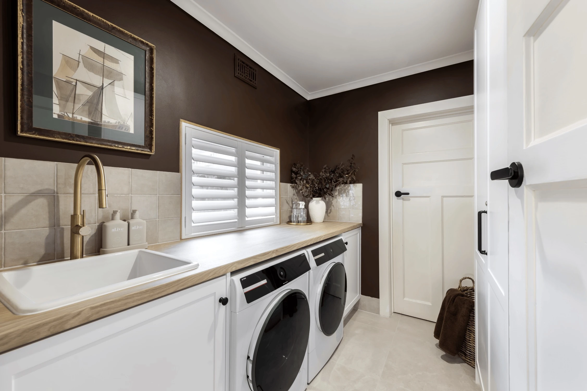 Modern laundry room with brown walls, white appliances, wooden countertop, sink with brass faucet, and framed ship artwork.