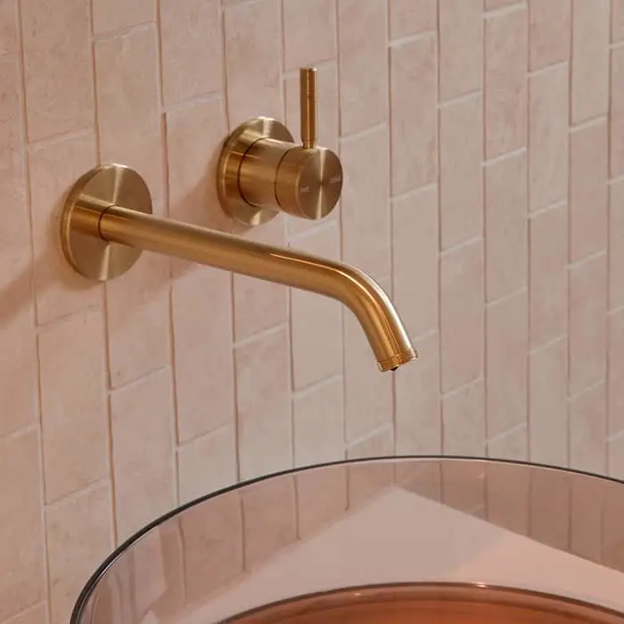 Brass wall-mounted bathroom faucet with separate control knob on pink tiled wall above a sink basin.