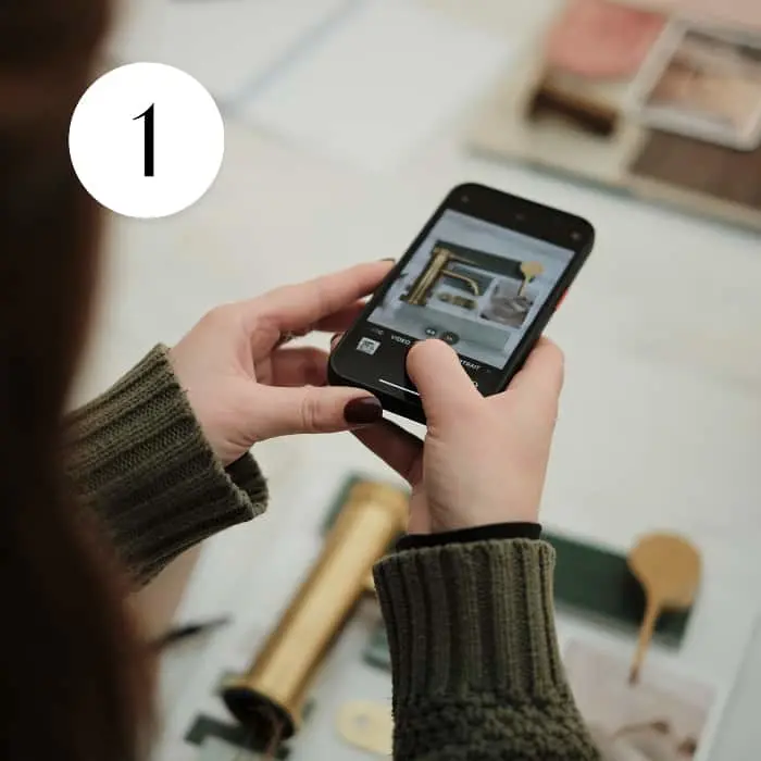 Person in a green sweater holding a smartphone, photographing design samples on a table. Number 1 in a white circle top left.