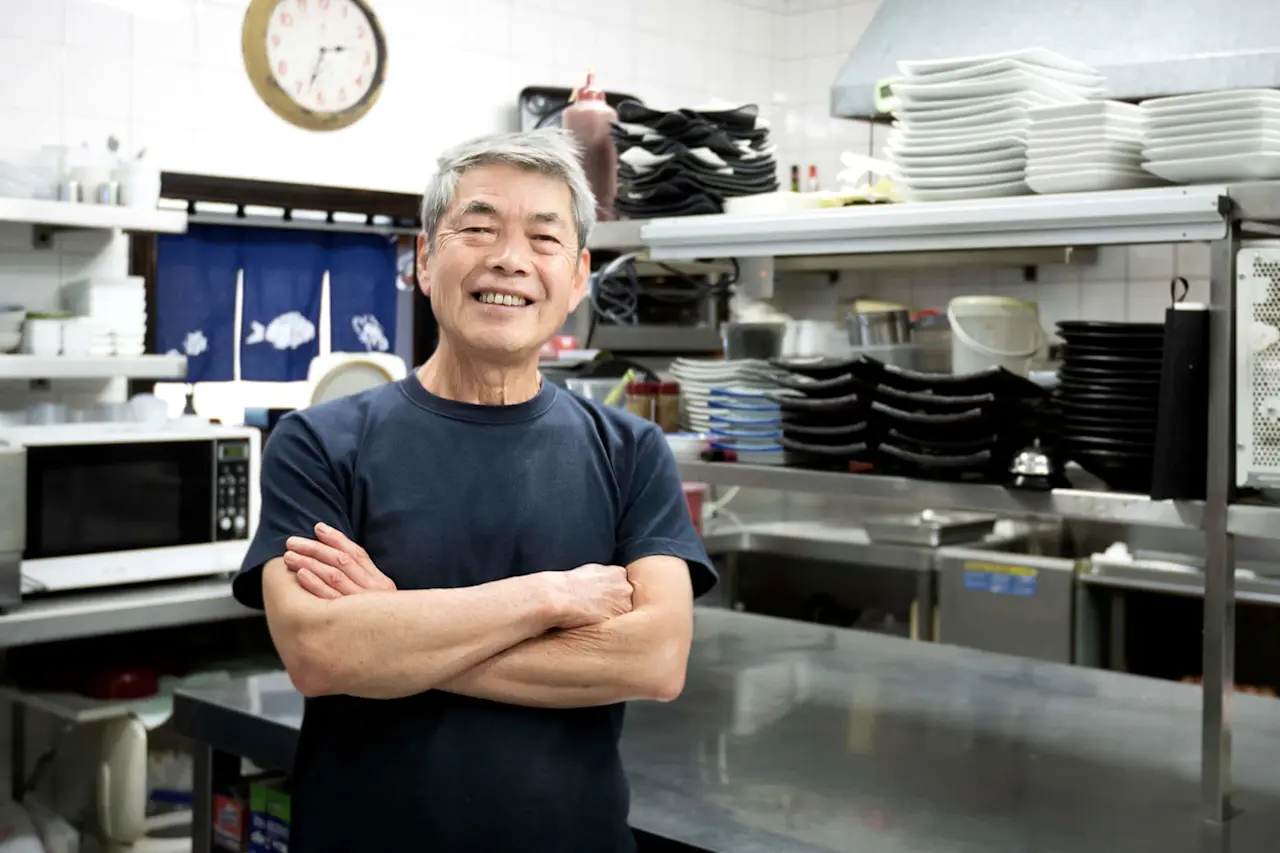 Smiling older man with arms crossed in restaurant kitchen with stacked dishes, blue curtain, and wall clock visible.