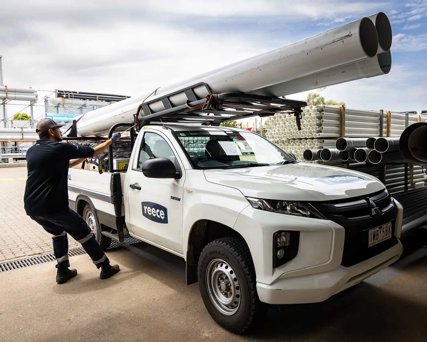 Worker loading large white pipes onto a Mitsubishi pickup truck with Reece branding at a supply yard.