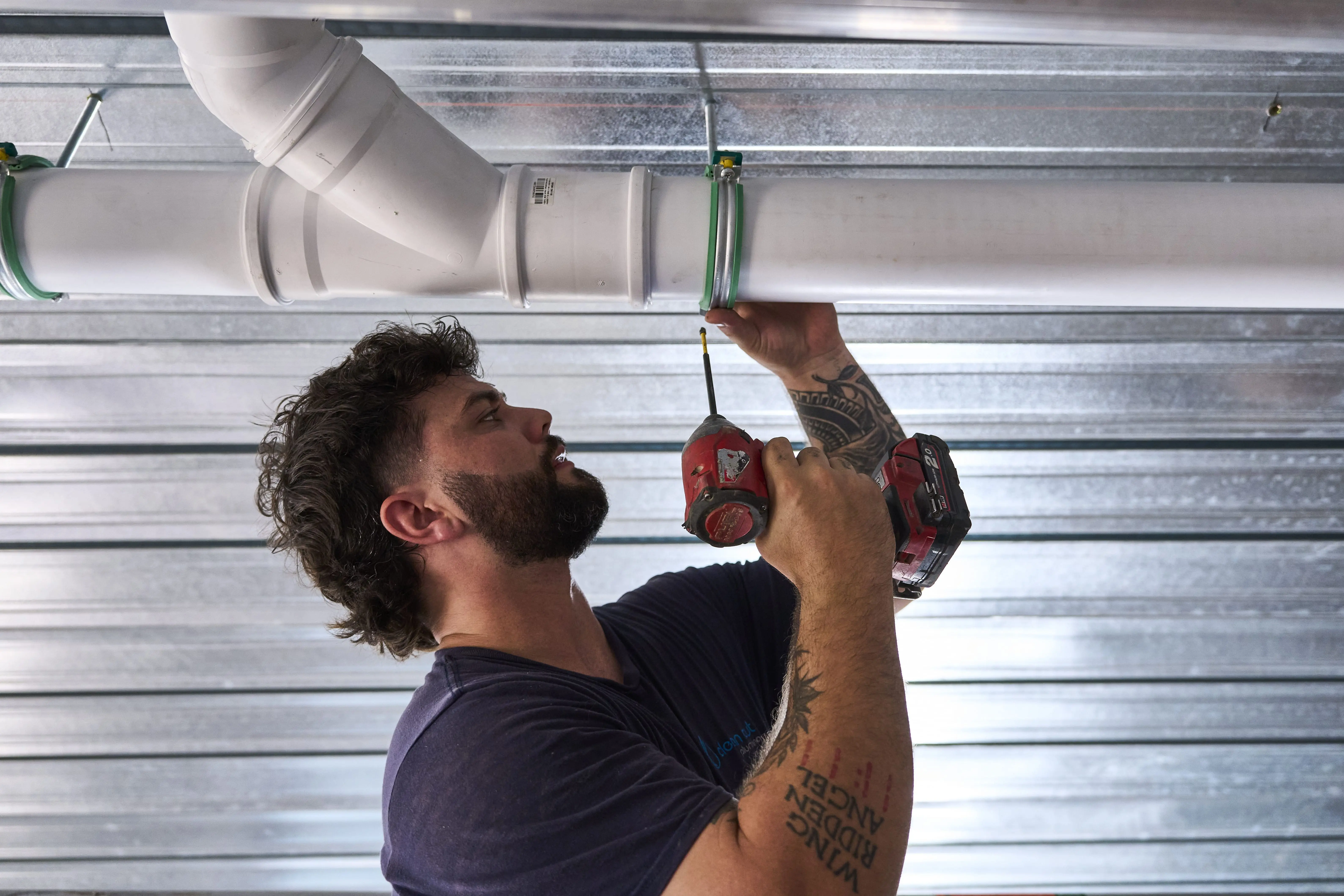 Plumber using red power drill to install white PVC pipe with green clamps on metal ceiling.