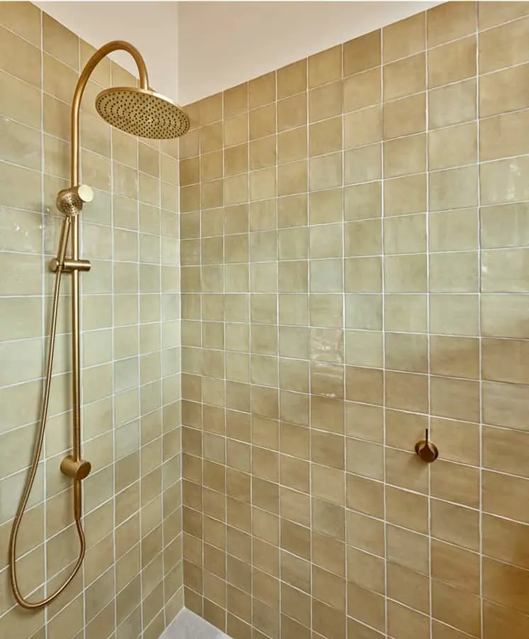 Modern shower with brass rainfall showerhead and handheld attachment against beige tiled walls.