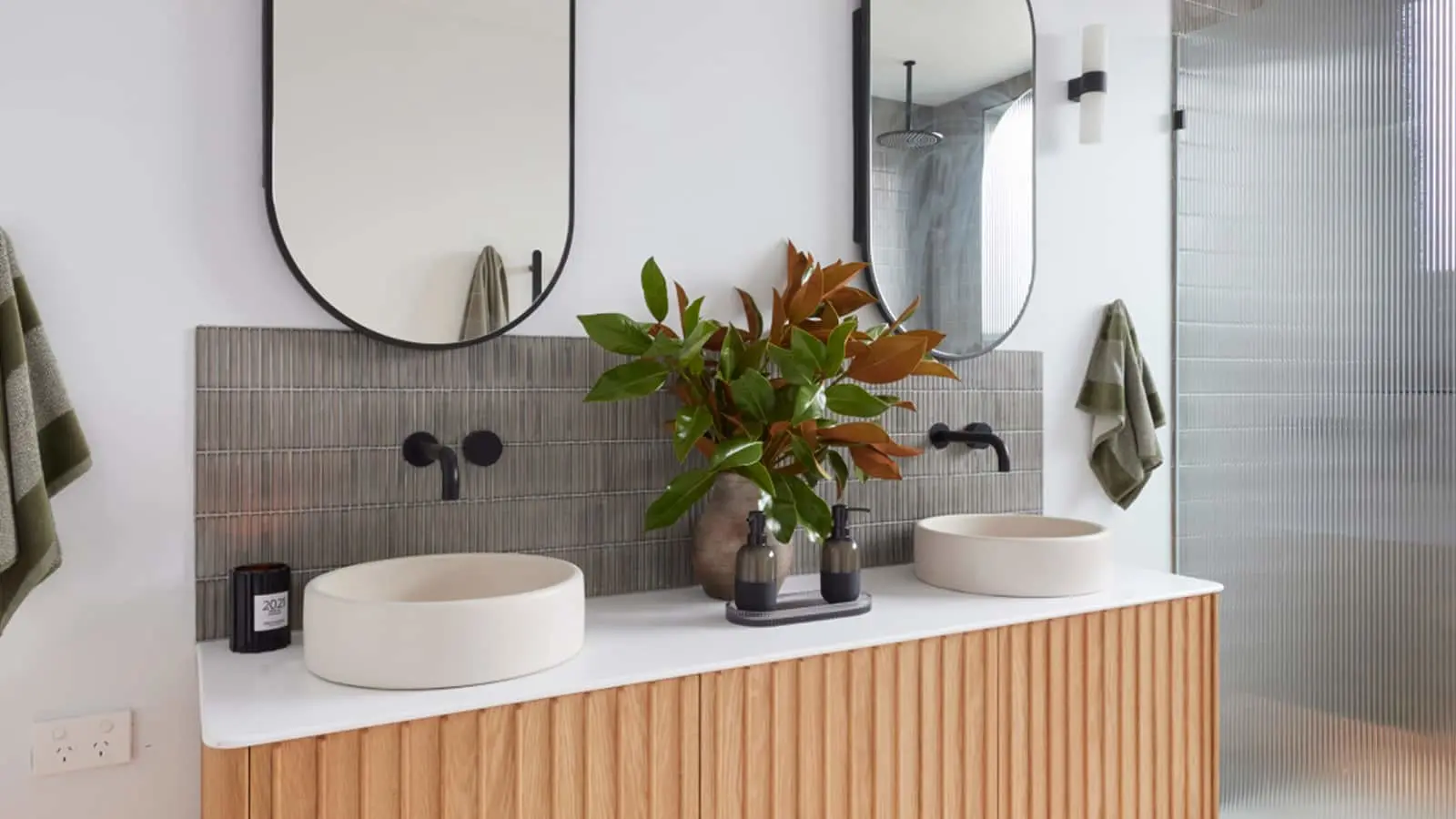 Modern bathroom with double vessel sinks, wooden vanity, round mirrors, and potted plant on white countertop.