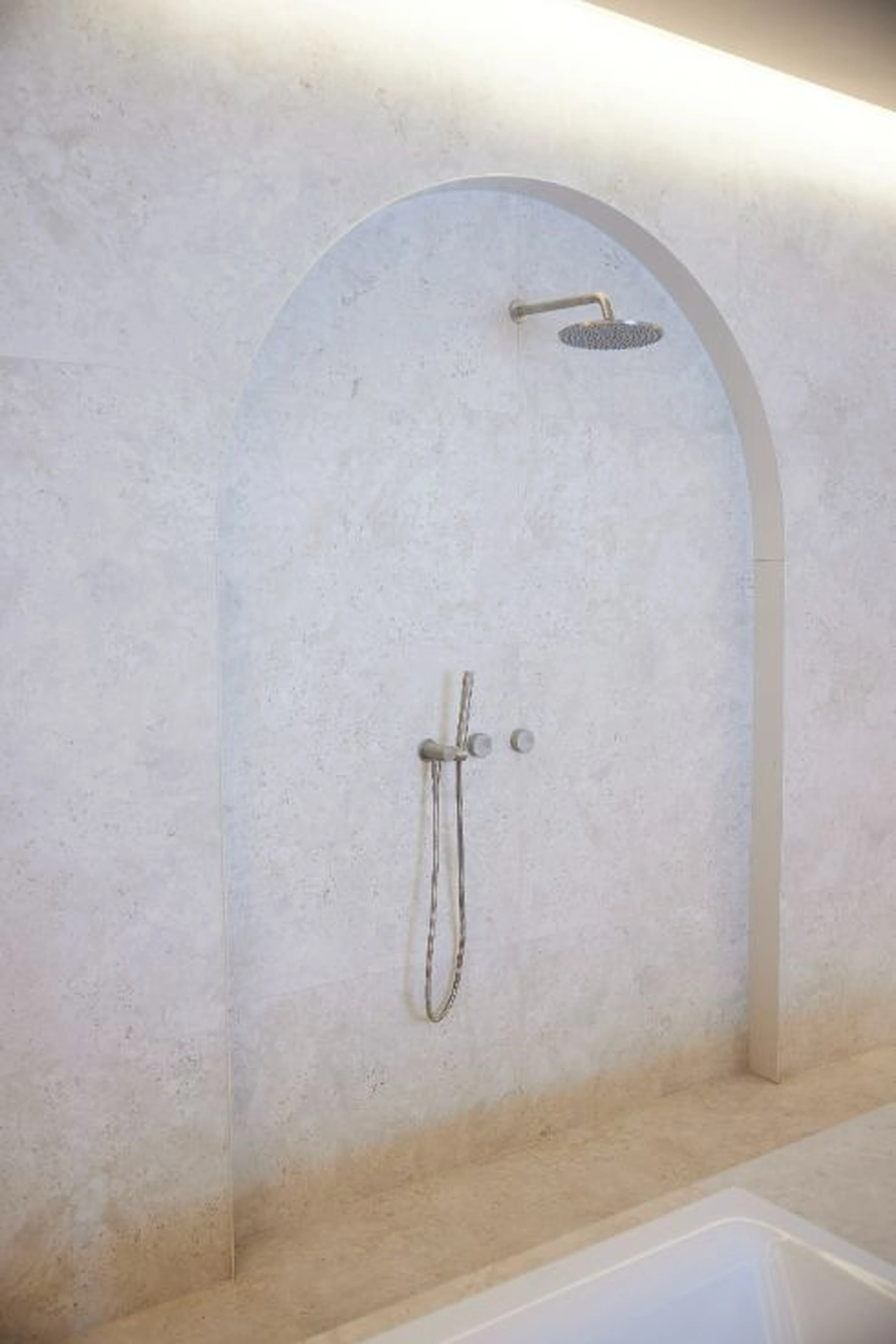 Minimalist shower with arched alcove, light stone walls, rainfall showerhead and handheld sprayer in brushed metal finish.