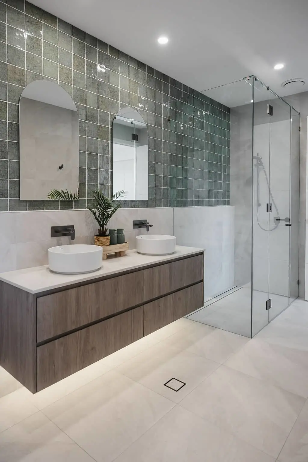 Modern bathroom with green tiled wall, wooden vanity with dual white vessel sinks, and glass shower enclosure.