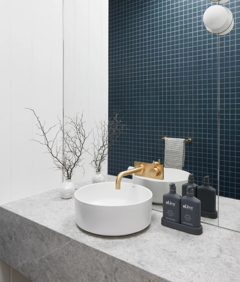 Modern bathroom with white vessel sink, brass faucet, blue tile wall, marble countertop, and black toiletry bottles.