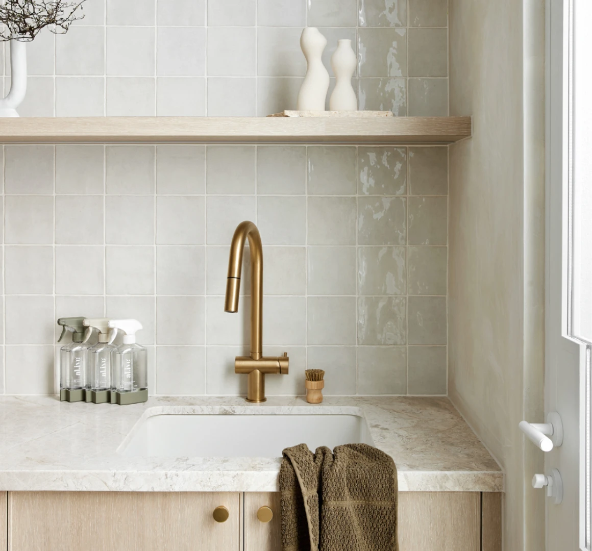 Modern kitchen sink with brass faucet, light tile backsplash, wooden shelf with decorative vases, and cleaning bottles.