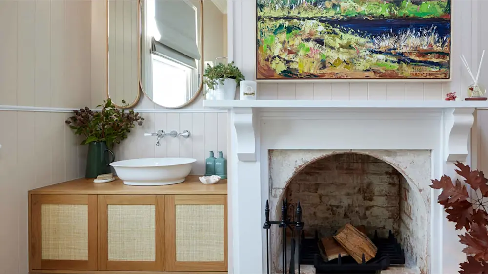 Elegant bathroom with vessel sink on wooden vanity, round mirror, white fireplace, and colorful landscape painting above.