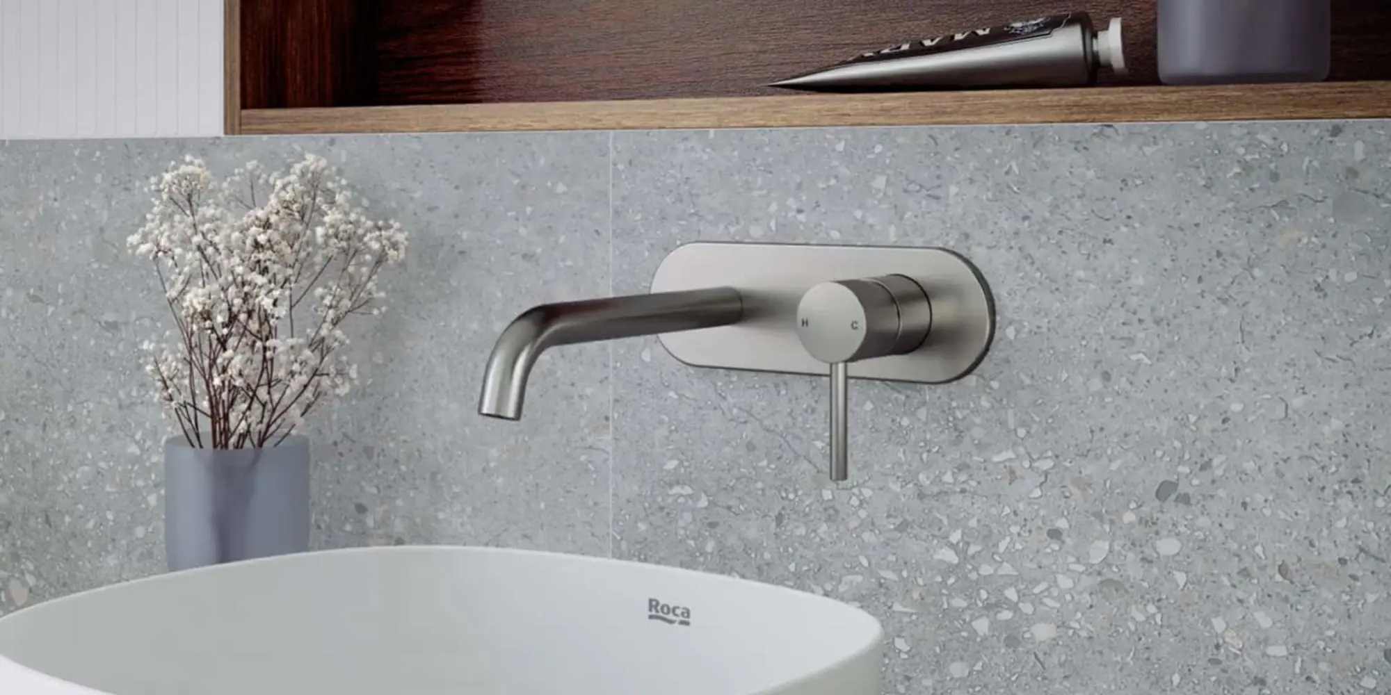 Modern bathroom with wall-mounted stainless steel faucet, white Roca sink, gray terrazzo wall, and small dried flowers.
