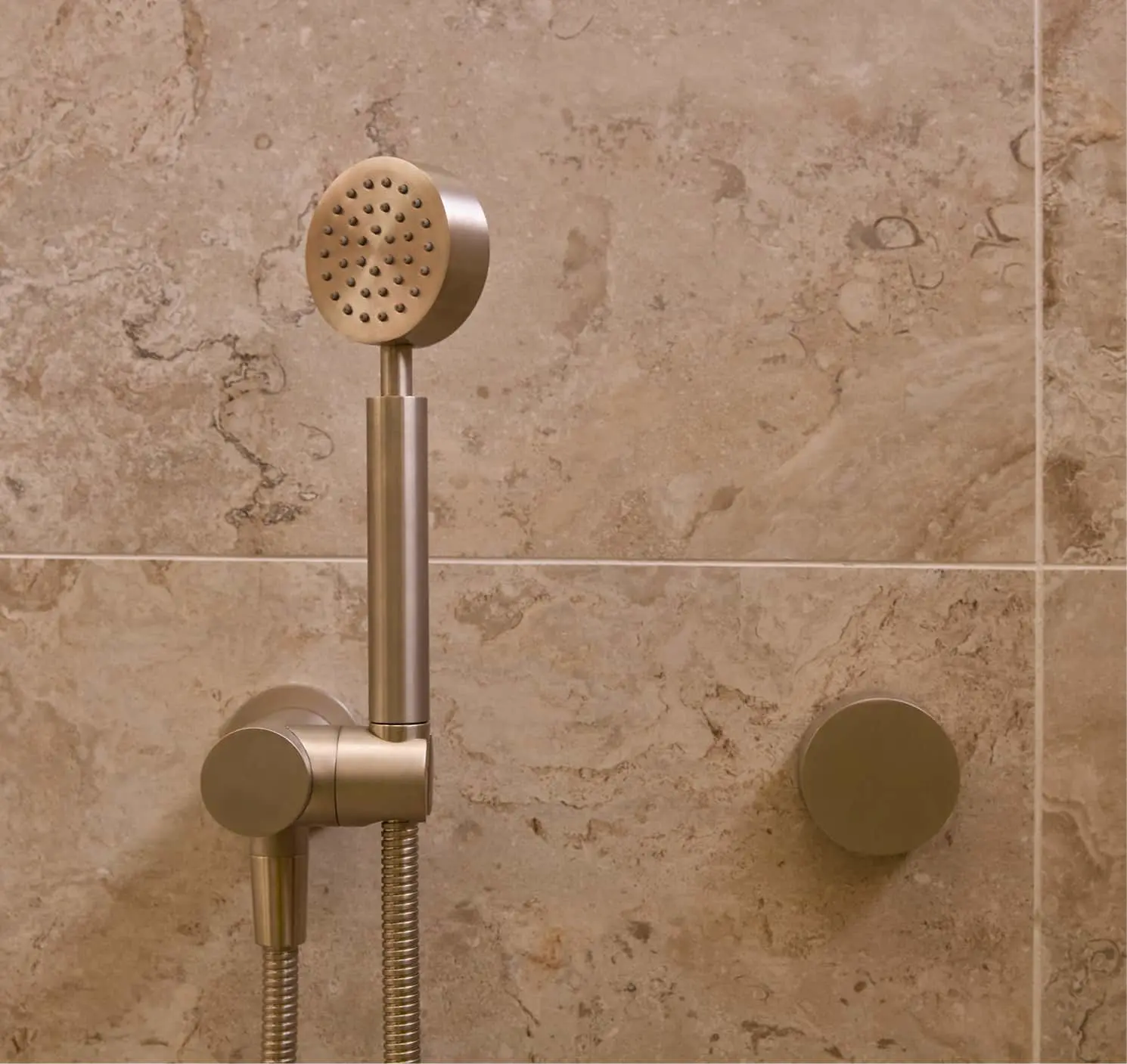 Brushed gold handheld shower head with holder and control knob mounted on beige tile wall.