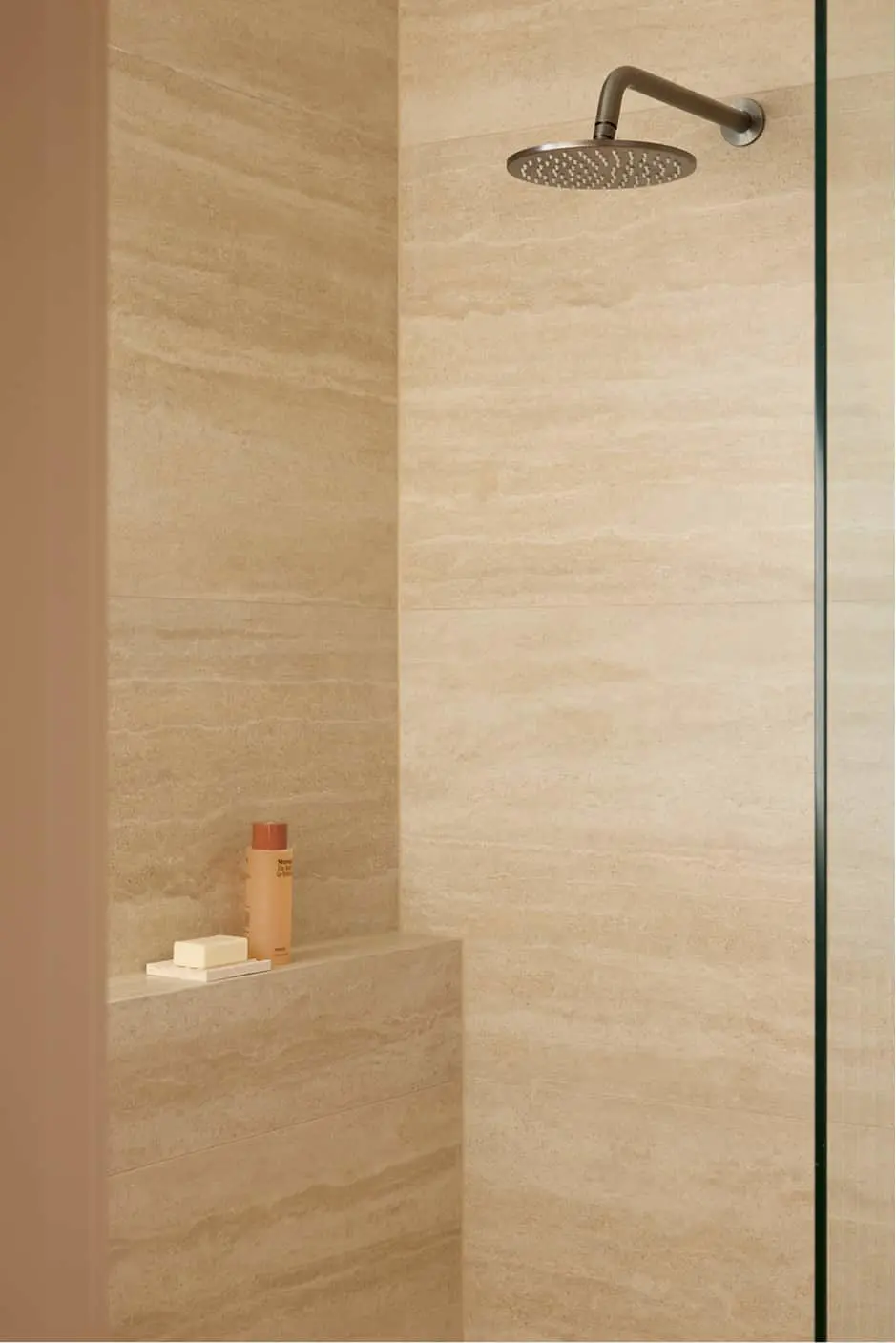 Beige tiled shower with gunmetal rainfall showerhead and glass partition, featuring a built-in shelf with toiletries.