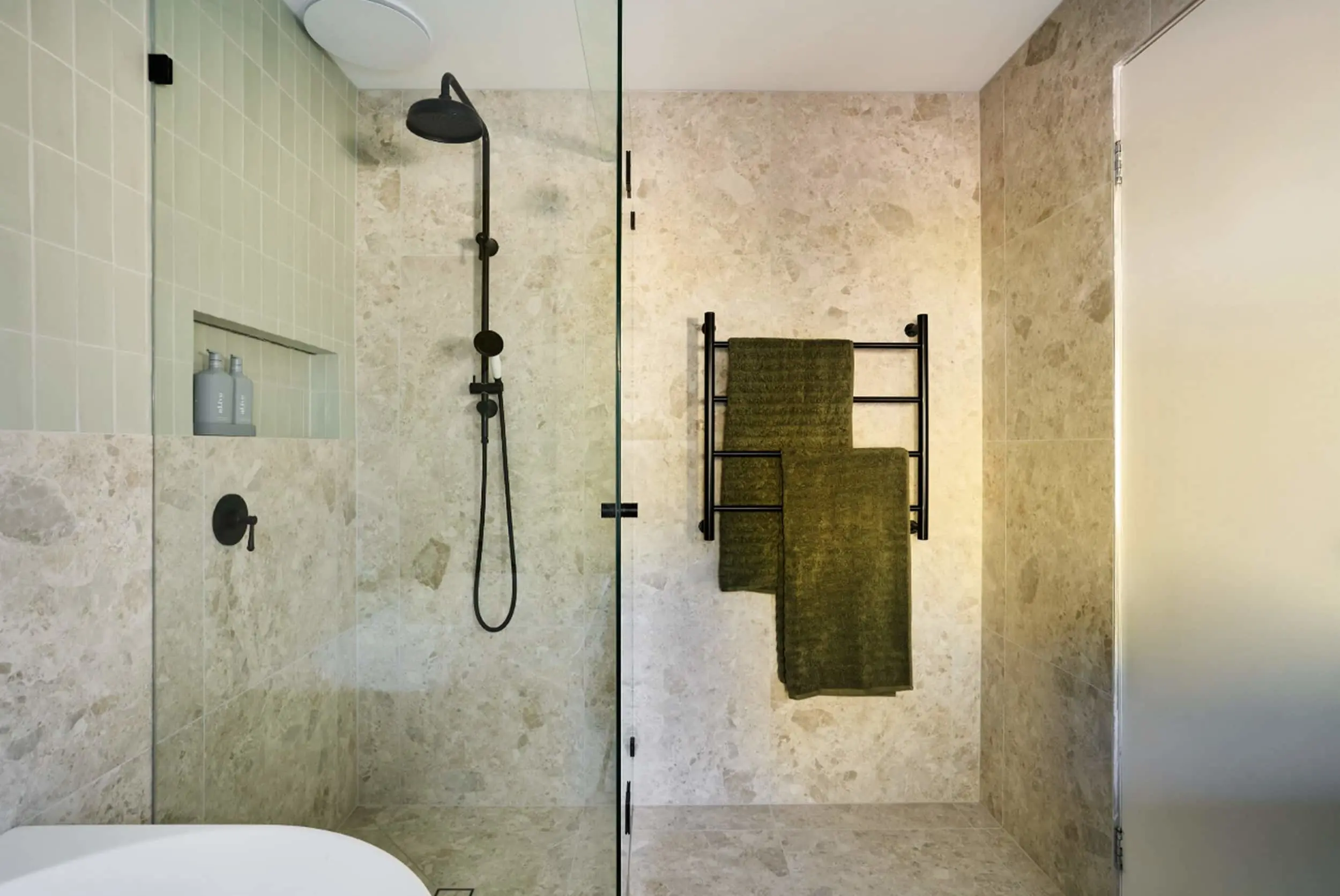 Modern bathroom with stone tile walls, black shower fixtures, glass door, and dark green towels on a wall-mounted rack.