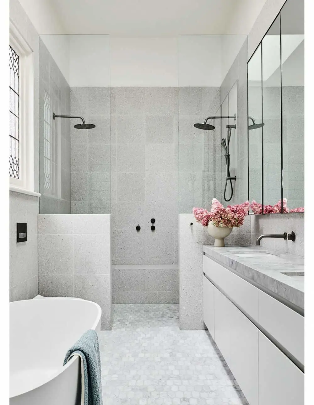 Modern bathroom with gray tile walls, walk-in shower, white vanity, freestanding tub, and pink flowers in a vase.