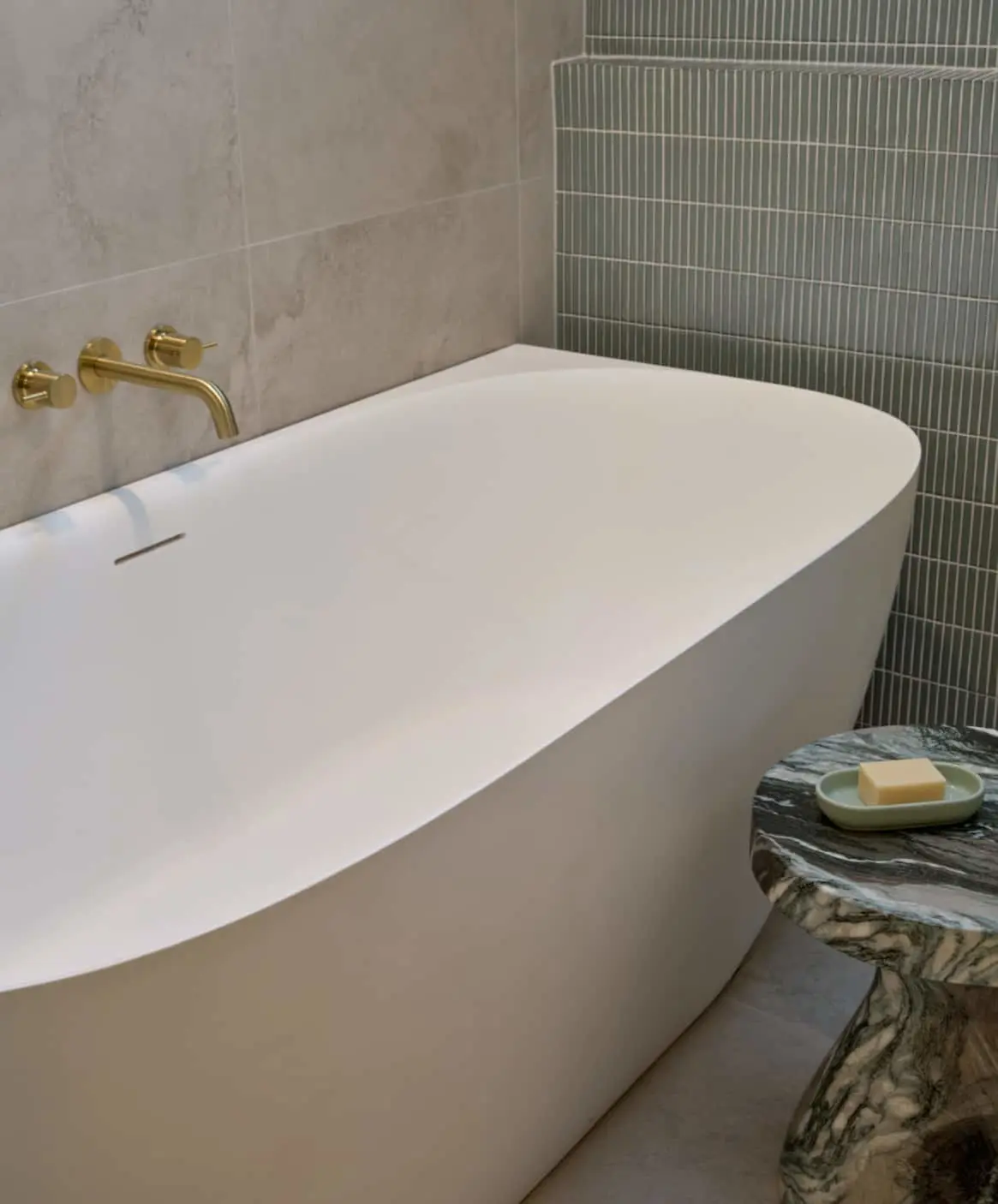 Modern white bathtub with brass wall-mounted faucet, green tiled wall, and marble side table holding soap dish.