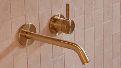 Brushed gold wall-mounted bathroom faucet with curved spout and separate control knob against pink tiled wall.