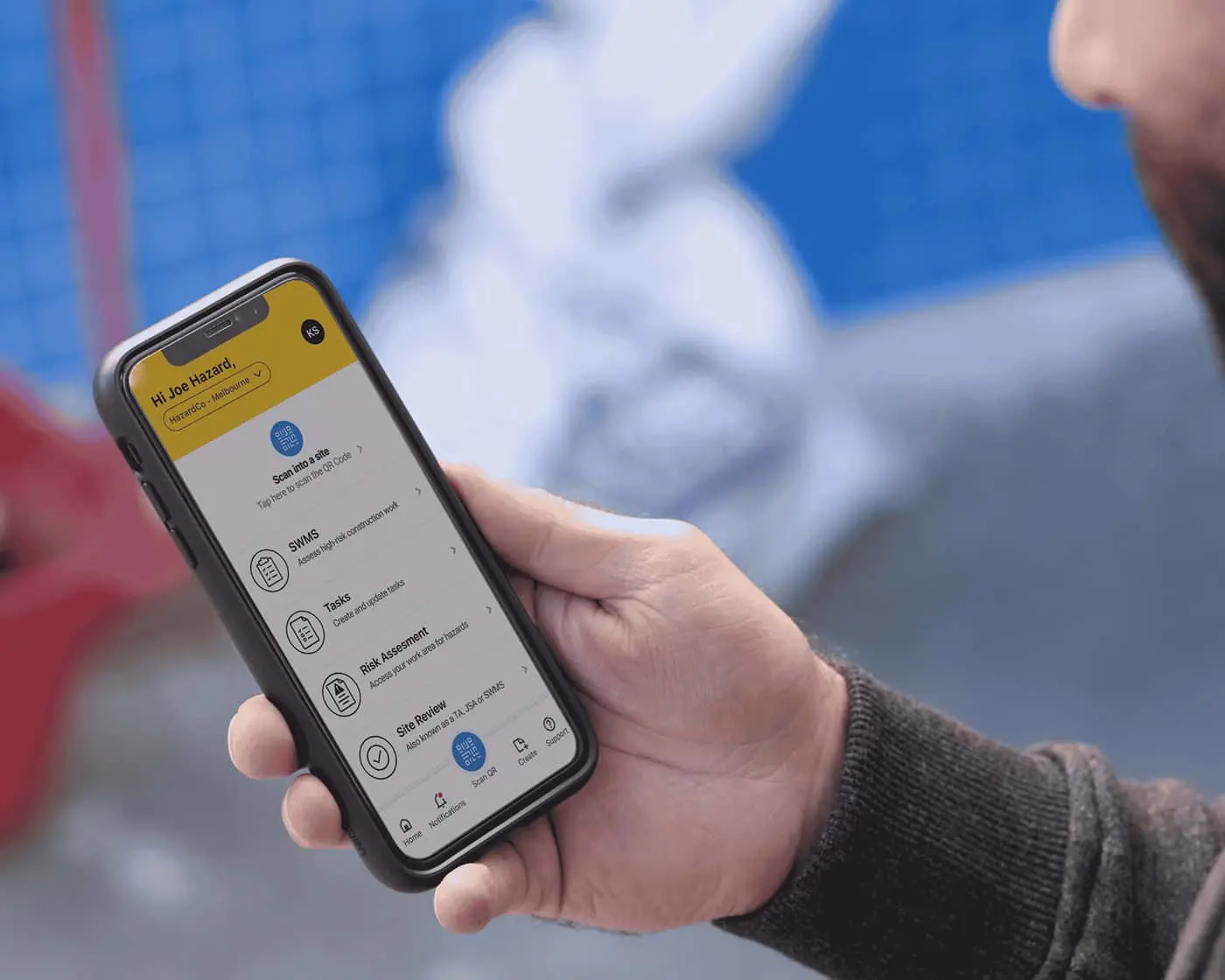 Person holding smartphone displaying safety app with SWMS, Tasks, and Risk Assessment options against blurred blue background.