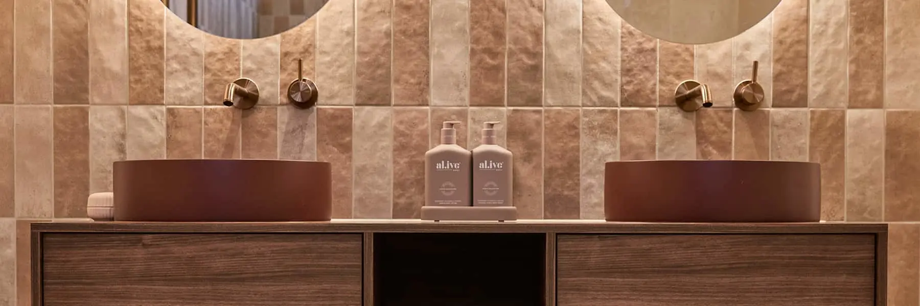 Modern bathroom with dual maroon vessel sinks, brass fixtures, beige tiled wall, and wooden vanity.