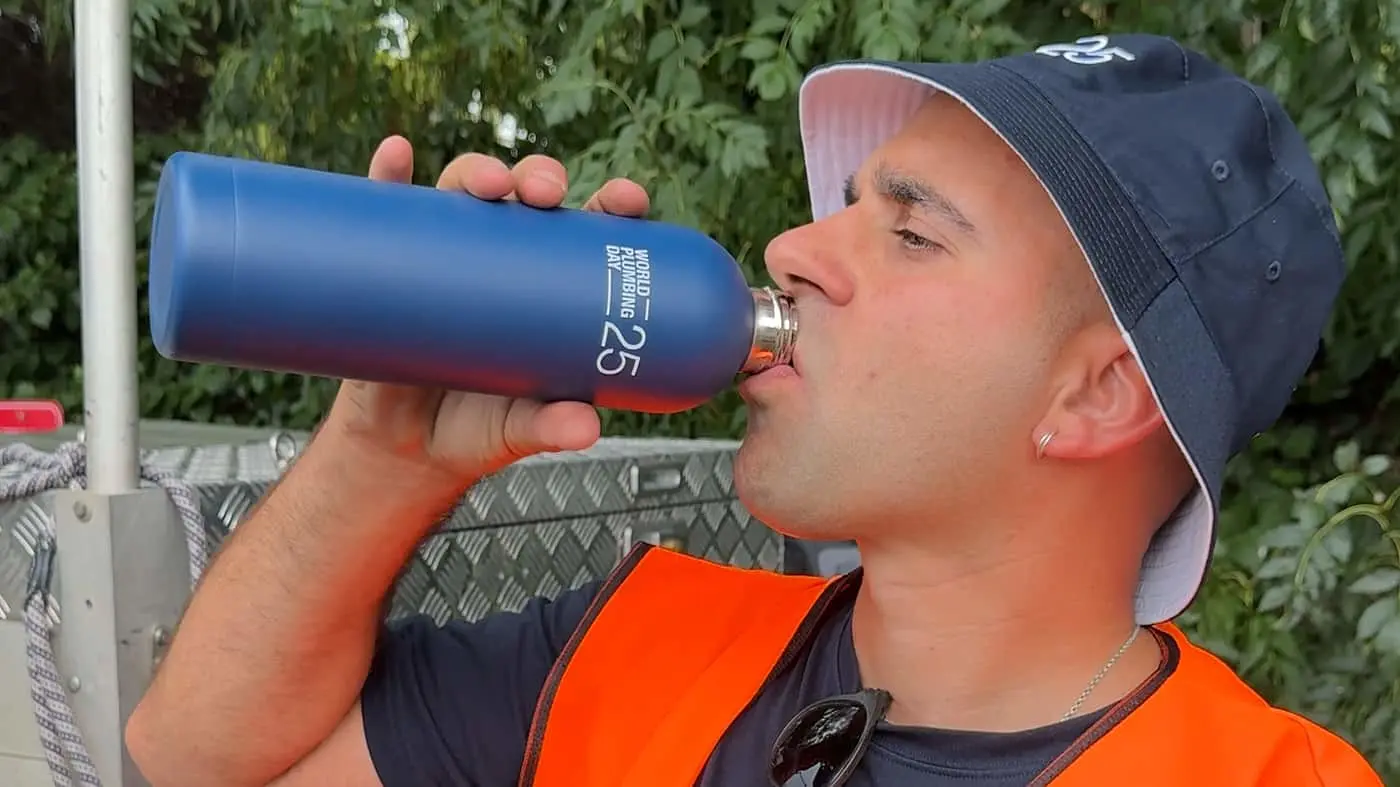 Person in navy cap and orange safety vest drinking from blue water bottle outdoors against green foliage.