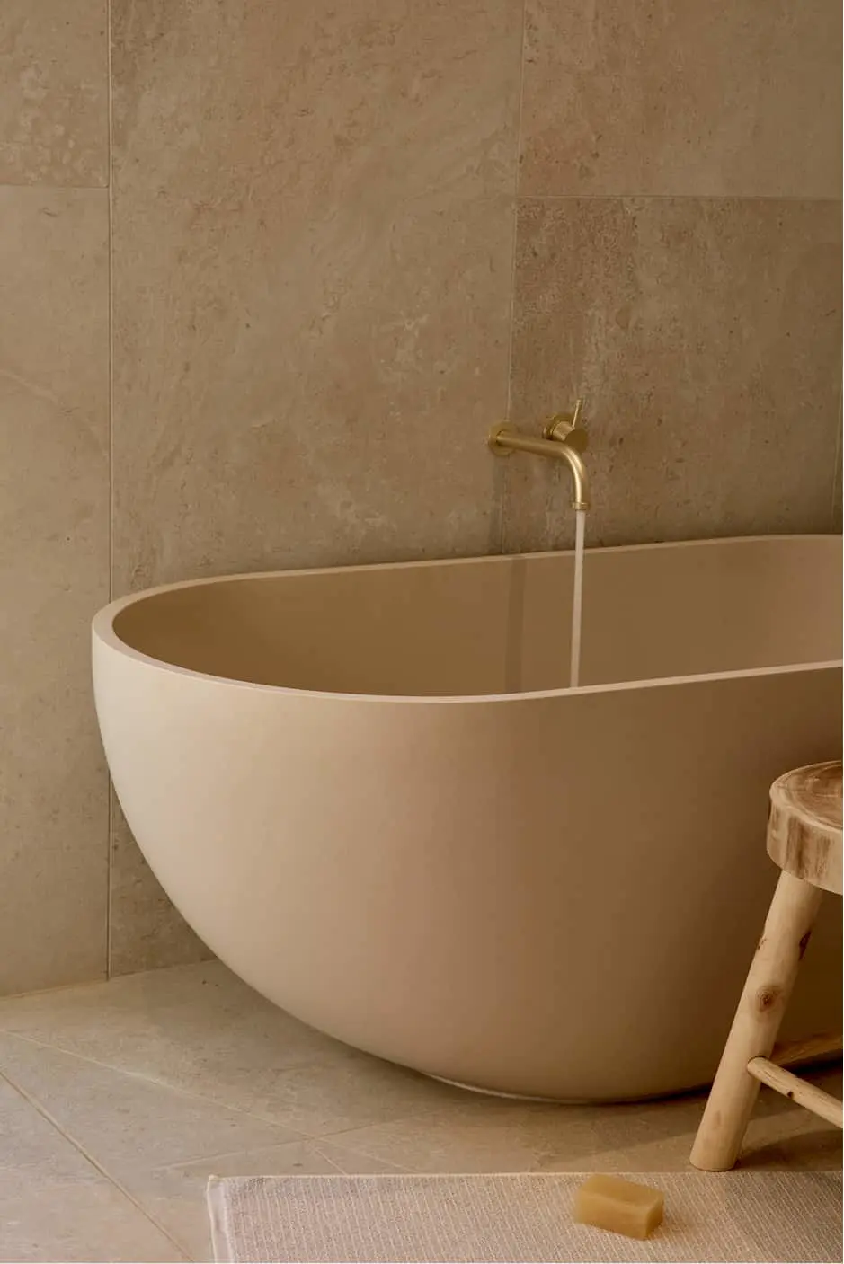 Beige freestanding oval bathtub filling with water from a brass wall-mounted faucet, beside a wooden stool, on stone tiles.