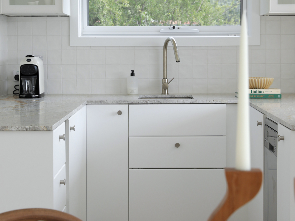 Modern white kitchen with marble countertops, stainless steel sink, coffee maker, and window overlooking greenery.