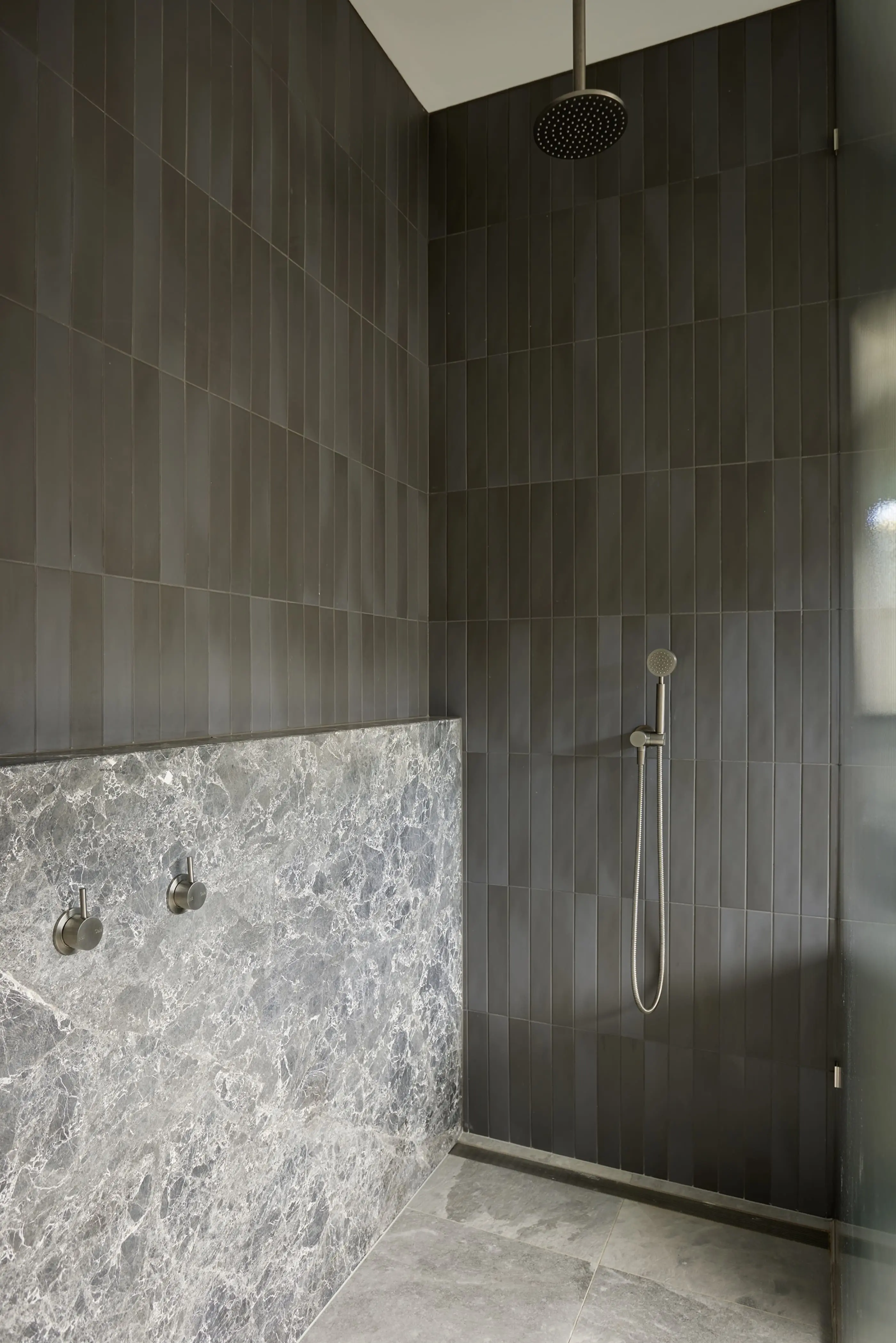 Modern shower with dark vertical tiles, marble lower wall, ceiling-mounted rainfall showerhead and handheld sprayer.