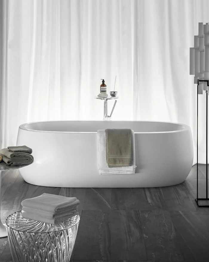 White oval freestanding bathtub on dark gray floor with towels draped over the side, a small tray with toiletries, and white curtains behind.