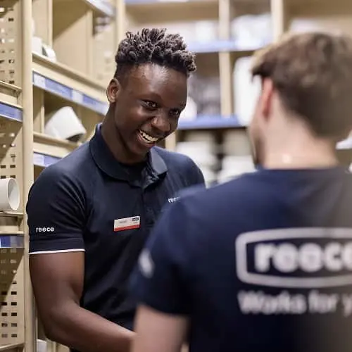 Two colleagues in navy blue Reece uniforms having a conversation in what appears to be a warehouse setting.